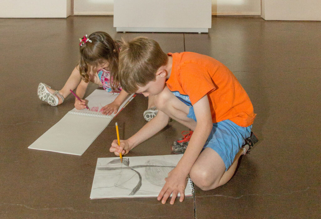 Two children are sitting on the foreground of this photograph. They are holding pencils and intently sketching abstract art.