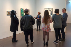 A view in a gallery of a female guide giving a tour to a group of five young people who are laughing and listening.