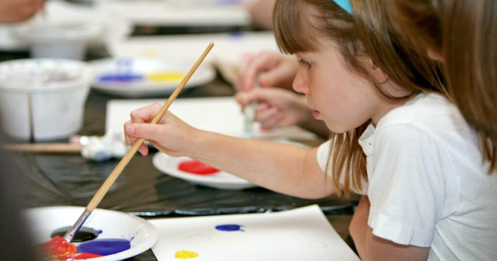 This photograph shows the side profile of a child holding a long paint brush. The tip of the paint brush is dipped into red ink. Her long hair is held back by a blue headband.