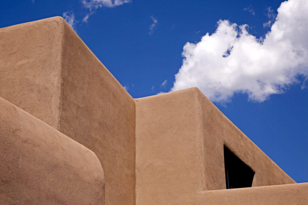 View of adobe style roof of the Georgia O'Keeffe Museum. Above the roof is bright blue sky with a cascading cloud.