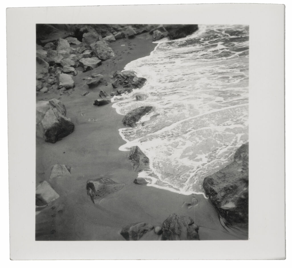 A black, and white photograph of sandy rocks in Hawaii. A wave washes up with foam.