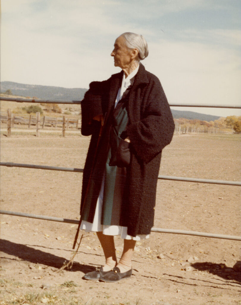A color photograph of O'Keeffe in New Mexico. She stands at a gate and behind her is a gravel landscape with mountains in the distance. She has her hands in the pocket of a large black coat. We see the side profile of her face and her white gray hair is pulled up at the nape of her neck.