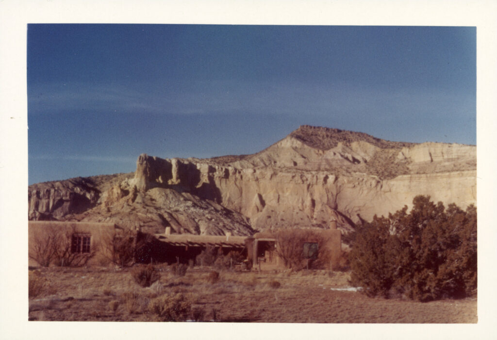 A sepia tone photographic snap shot of the outside of Georgia O'Keeffe's adobe home at Ghost Ranch. The adobe home blends in with the gravel and brush in the foreground. A white rocky mesa shoots out from behind the flat one story adobe home in contrast against the blue sky.