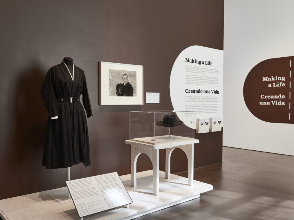 A photograph of an exhibition installation showcasing the work of artist Georgia O'Keeffe, titled "Georgia O'Keeffe: Making a Life." The photograph captures a section of the exhibition, with several of O'Keeffe's paintings, drawings, and photographs displayed on the walls.