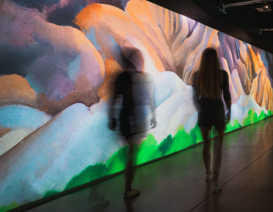 Shadows of two human figures slightly blurred as if in motion, they walk past a colorful wall that looks like a mountain landscape of with warm yellows in the background and cool blues and green in the foreground.