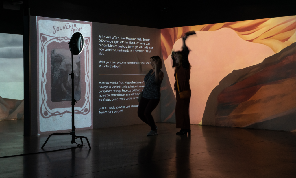 A photograph of the Photo Booth at Music for the Eyes in Albuquerque. A selfie stick station is in the foreground and a pink canyon oil painting by Georgia O'Keeffe is in the bankground.