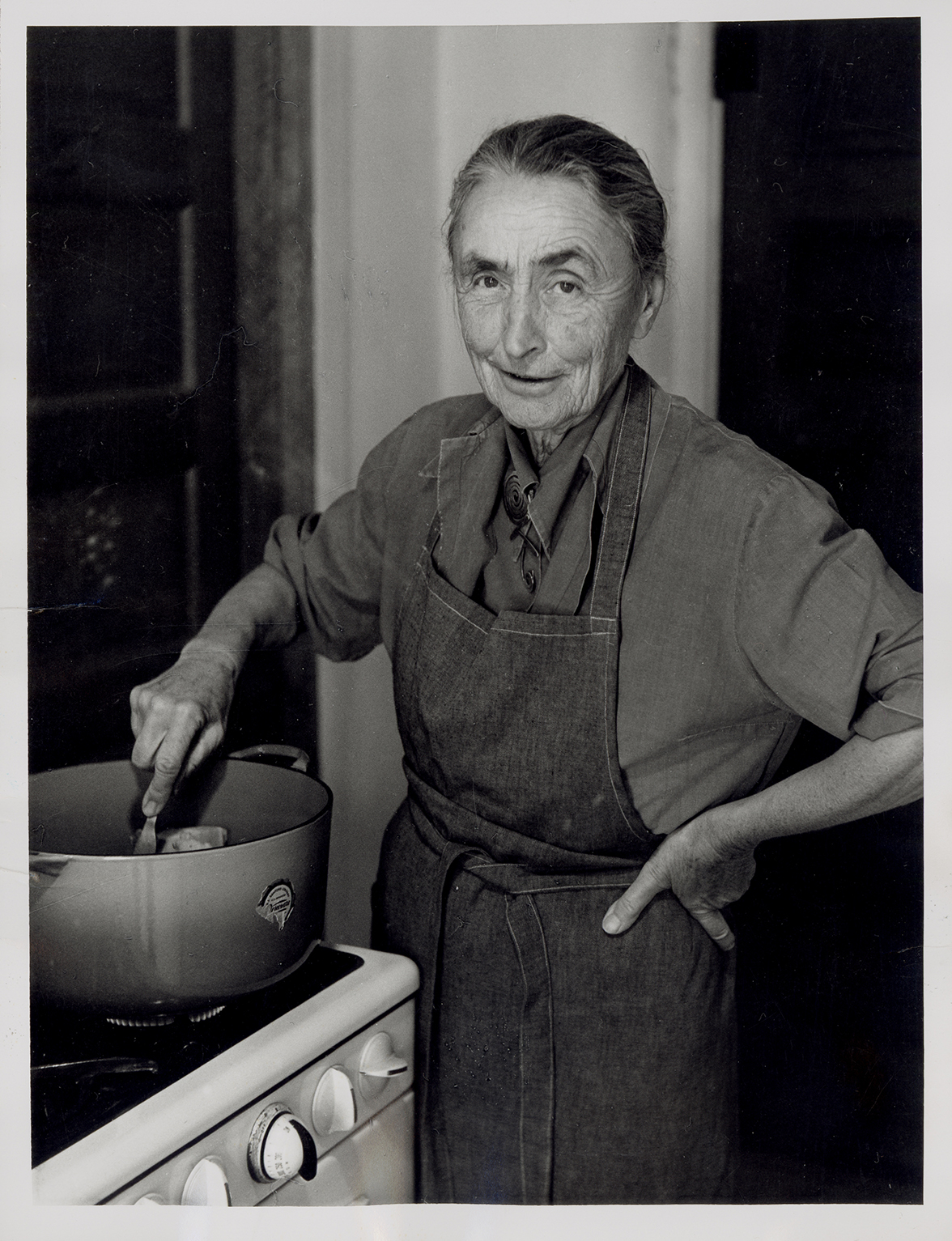 Georgia O'Keeffe at a stove. She wears a denim apron and an open mouth smirk. Her hair is pulled back and she holds a wooden spoon and stirs something on the stove.