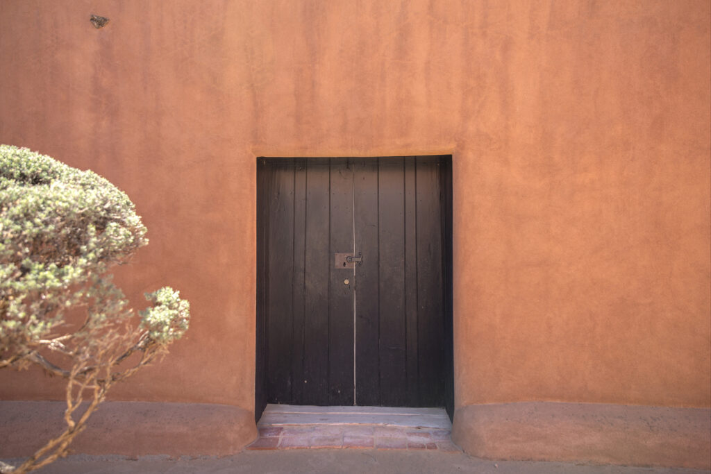 This photograph features the outside patio and door at Georgia O'Keeffe's Abiquiu Home and Studio. Foliage is visible on the left side of the frame.