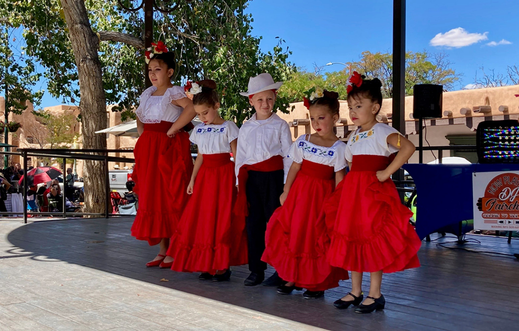 Photograph of five children in traditional folkloric dance dresses and clothing from Mexico standing in line on a stage.