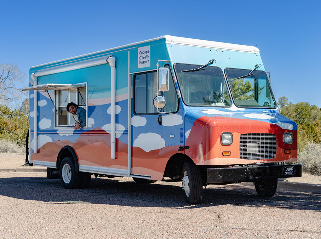 Photograph of the Art to G.O. Truck, a mobile creativity studio with motifs of desert landscapes and clouds on it. In the truck's window is a person peering out. Behind the truck are green trees and a blue sky.
