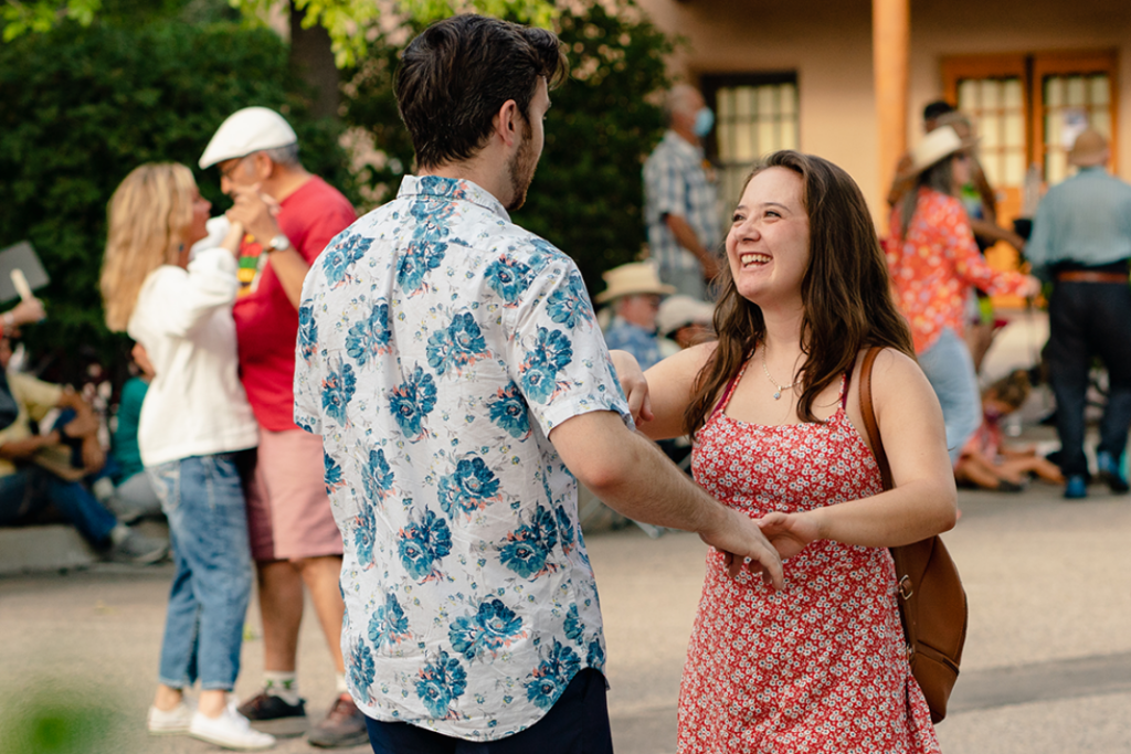 Photograph of two people dancing and smiling together holding hands. Behind them are trees, an adobe building and other couples dancing.