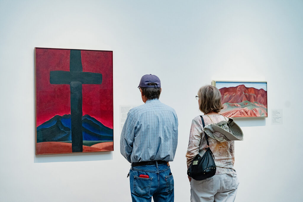 Photograph of two people, one in a baseball cap and one carrying a bag, looking at two paintings on a white gallery wall. The painting on the right is a horizontal landscape of dark reddish crimson clay hills. The painting on the left is a vertical painting of a large black cross set against a bright red sky with a dark mountain in the background.
