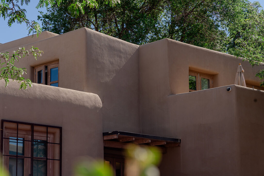 Exterior of an adobe building with two levels and several windows. The adobe building is framed by green trees and plants in the foreground and background.