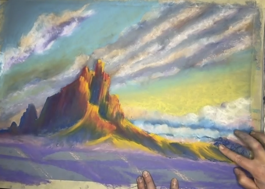 A pastel drawing depicting a vibrant desert landscape with a prominent red rock formation, a yellow and green sky, and wispy clouds. Two hands are visible, adding details to the artwork.