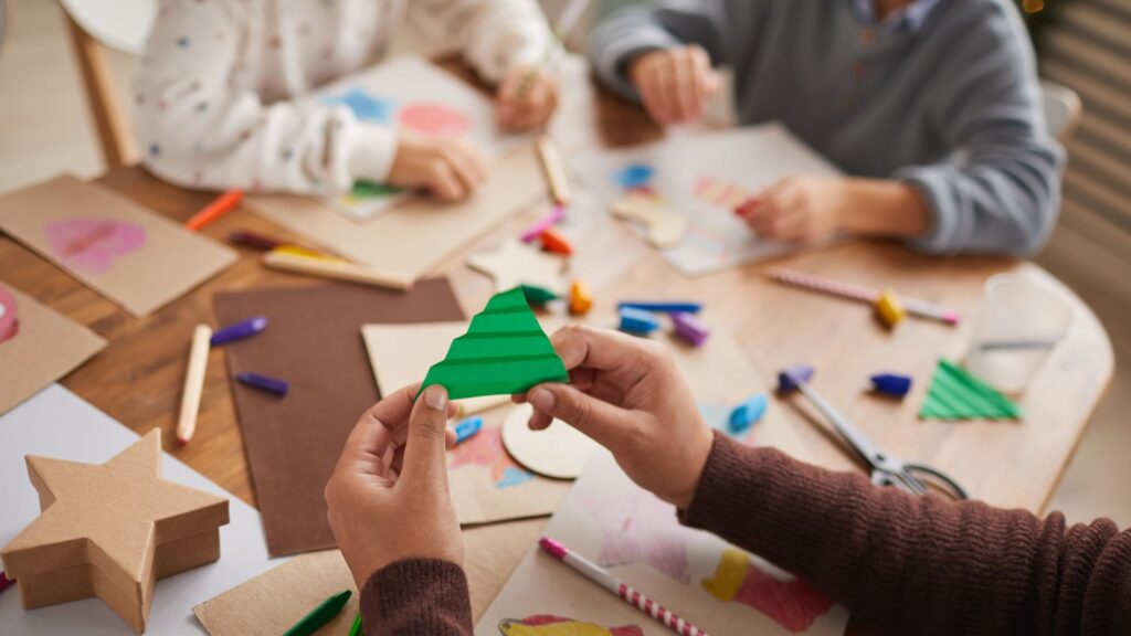 Photograph of a person's hands as they hold up apiece of folded green paper. In the background are two children's hands as they make collages.