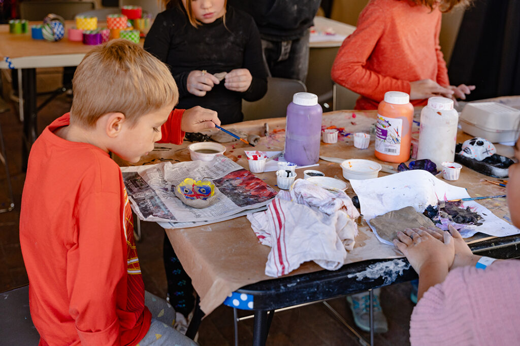Photograph of a group of young children painting clay bowls made by hand at a table covered with art supplies.