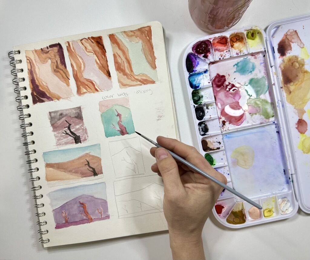 Photograph from above of a person's hands as they paint different small scenes on a notebook with colorful watercolors. To the right is a watercolor palette.