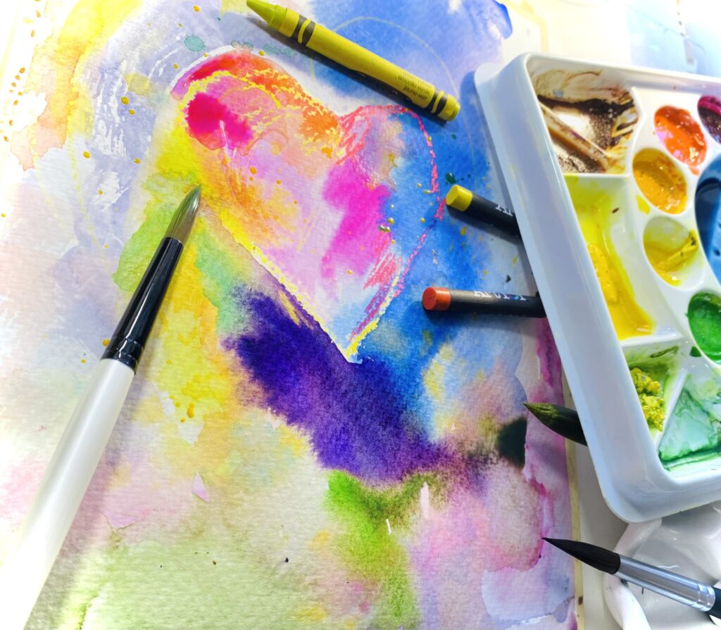 Photograph of a heart and colorful hues of watercolor paint on a canvas. On the canvas are paintbrushes, crayons and a watercolor palette.