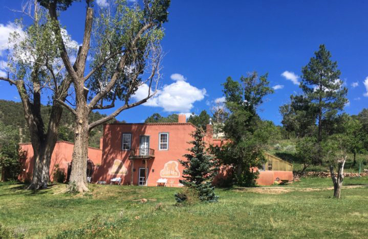 Photograph of an adobe home in pink hues surrounded by green trees. Behind the home is a clear blue sky