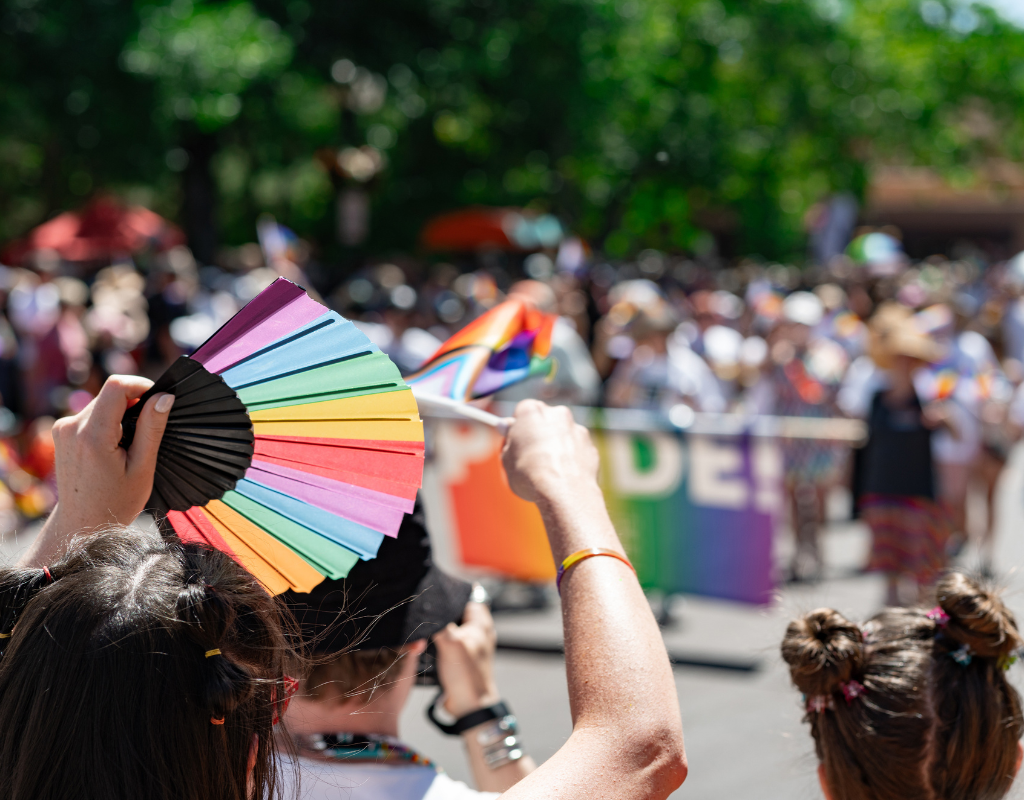 Photograph of hands waving a rainbow fan and a Pride flag in front of a crowd of people celebrating Pride. Behind are green trees.