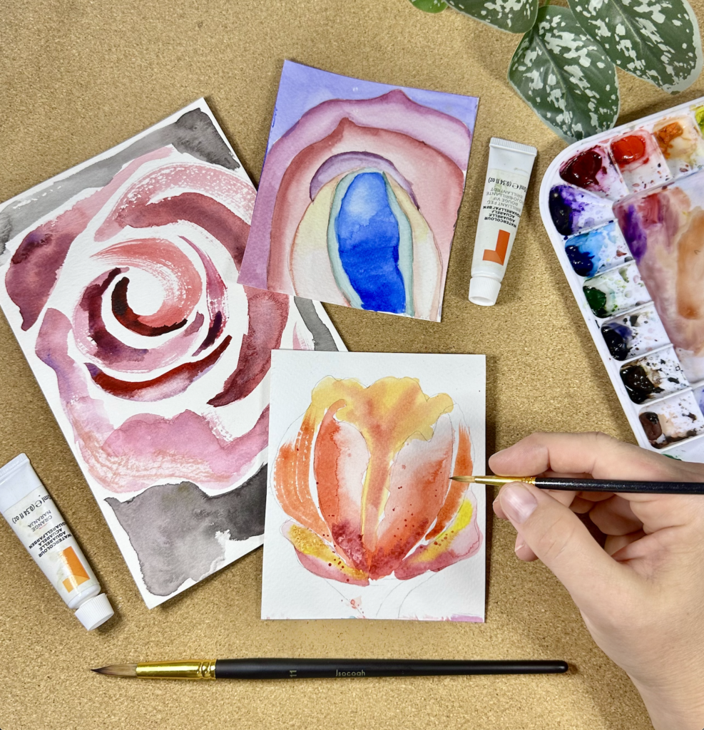 Photograph taken from above of an artists hand as they paint colorful abstract florals on small canvases using a paintbrush and a nearby palette.
