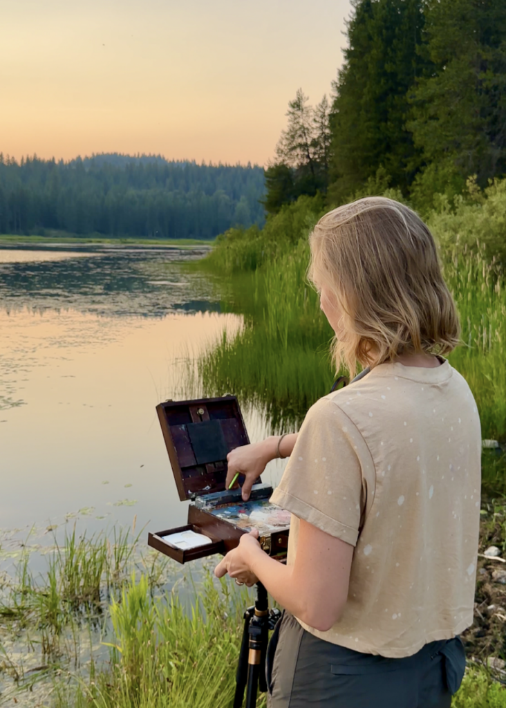 Photograph of a person with short hair standing at the edge of water with a small painting case and palette in front of them. in the background are green grasses, trees, and mountains surrounding the water.