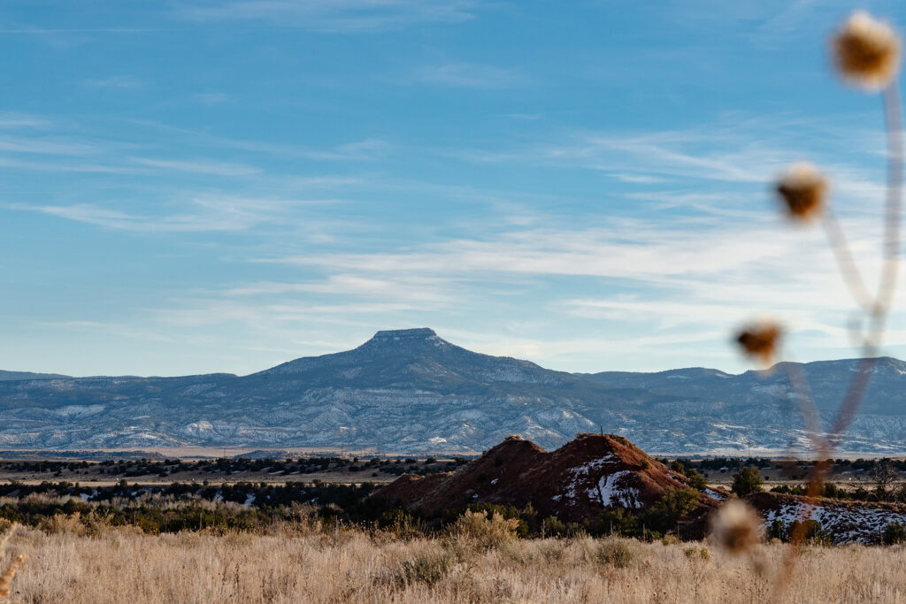 Photograph of Cerro Pedernal, or Tsee p'in, in the Tewa language. A flat-topped mountain with a vast desert landscape below. Behind the mountain is a blue sky and low clouds. To the right of the frame is a blurry plant.