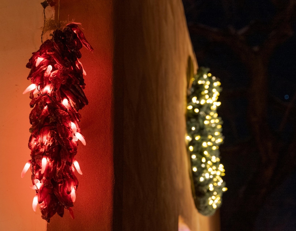 An adobe wall on which is a red ristra and a green wreath. Both are covered in lights. Behind the building is a dark sky.