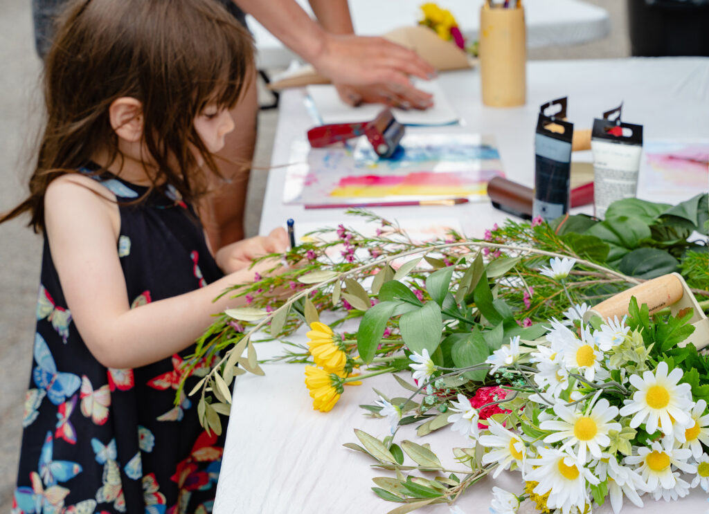 Photograph of a young child making an art piece on a table that is covered in art supplies and flowers.