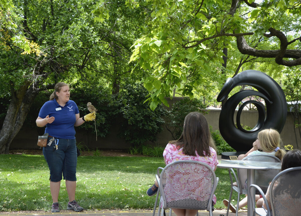 A person holding a small owl in front of a group of people sitting at tables outdoors. Behind the owl handler are green trees and an abstract sculpture