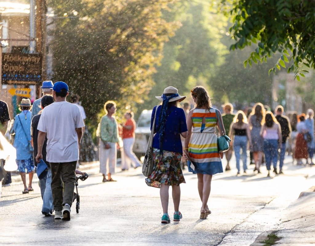 A crowd of people walk down a street in the Summer sunlight. Green trees line the street.