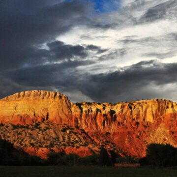 A beautiful photograph of the Red Rock Cliffs at Ghost Ranch at Sunset. The Red Rock Cliffs are illuminated in golden hour light. The background is filled with darkened moody clouds.