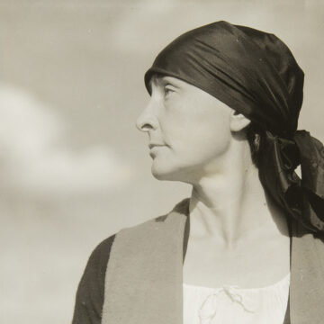 Half-length portrait of Georgia O'Keeffe with head in profile facing viewer left wearing a dark scarf/ turban tied behind her head; torso is frontal, neutral facial expression with eyes looking upward; she wears a white scoop-neck drawstring blouse under a dark sweater and vest; framed against a sky with clouds