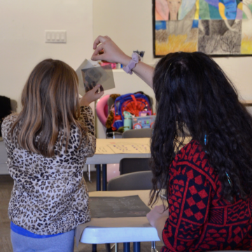 Photograph of a person and a child looking at photographic paper together. The child stands while the adult is sitting and they are each holding the photographic paper in the air with a hand.
