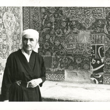 Black and white photograph of Georgia O’Keeffe wearing a dark wrap dress and a light scarf around her head at a Mosque. Behind her are the intricate tiles of the Mosque wall and to the left of the frame is gentleman passing through in front of the camera.