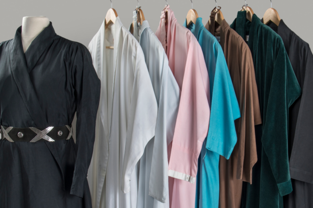 Eleven of O'Keeffe's dresses hang on a rack. Those on the left are dark shades of black and grey, and on the right, they range from white to light blue and pink. A black wrap dress with a belt is on a mannequin apart from the others on the left side of the frame, facing the camera.