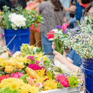 Image of a bunch of bouquets of flowers in containers and buckets on a table. In the background is barely visible a person arranging the flowers