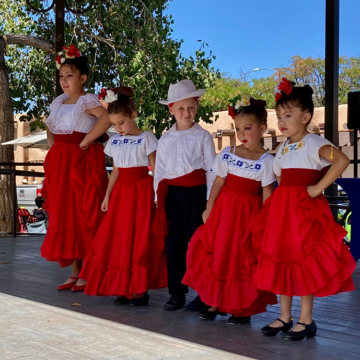 Photograph of five children in traditional folkloric dance dresses and clothing from Mexico standing in line on a stage.