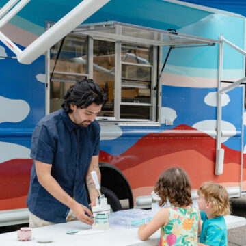 Photograph of a person standing over a table demonstrating making buttons across from two smaller children. Behind them is the Art to G.O. truck with clouds motifs on in.