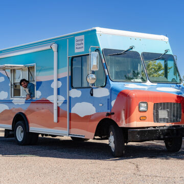 Photograph of the Art to G.O. Truck, a mobile creativity studio with motifs of desert landscapes and clouds on it. In the truck's window is a person peering out. Behind the truck are green trees and a blue sky.