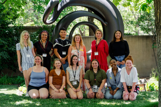 Photograph of a group of young people standing in a line in front of a dark cast of O'Keeffe's large 'Abstraction' sculpture. They are all looking at the camera and smiling. Around them are green trees, bushes, and a lush lawn.