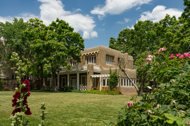 Photograph of the exterior of a two-story adobe building with a green lawn in front and a blue sky behind. The building is surrounded by trees and pink flowers occupy the foreground.