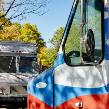Photograph of the front of a grey mobile art gallery in the background and the front of the Art to G.O. truck in the foreground. Behind both trucks are leafy trees turning gold.