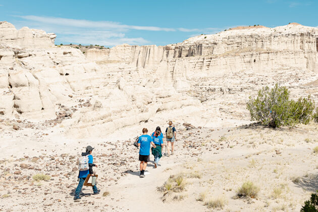 Photograph of a group of young students, many wearing blue shirts, backpacks, and hats, walking through the desert landscape known as the Plaza Blanca, or the White Place. They are small in the frame and the desert landscape extends all around them. The upper portion of the frame shoes a clear blue sky.