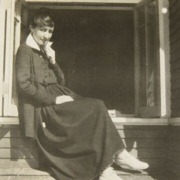 A black and white photograph of a woman sitting on a window ledge staring forward. She is wearing a black dress down to her ankles with a white collar. Her legs are crossed. The house is wooden clapboard.