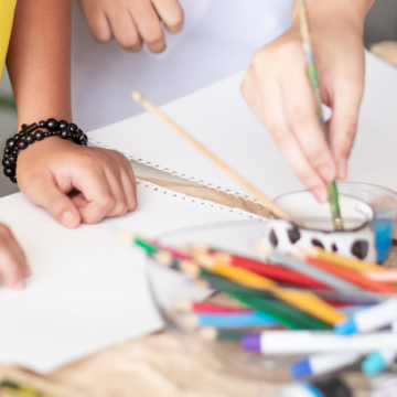 Close-up photograph of two young children's hands on a table as they paint and an adult's hands poke into view.