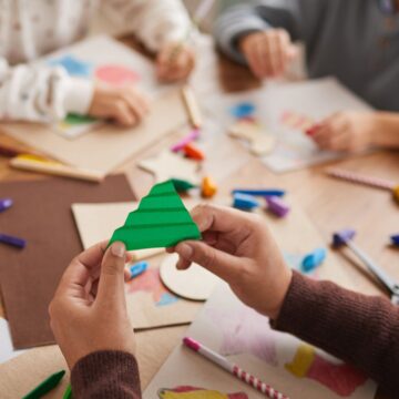 Photograph of a person's hands as they hold up apiece of folded green paper. In the background are two children's hands as they make collages.
