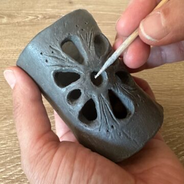 Close-up photograph of hands holding a small tubular lantern made of dark clay as they shape motifs and windows with a sharp and long wooden tool.