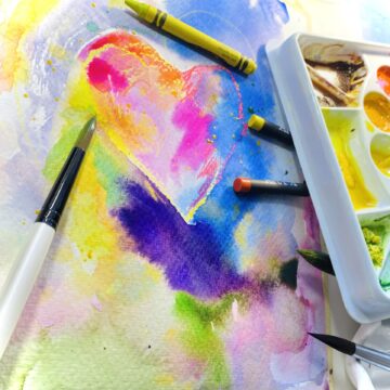 Photograph of a heart and colorful hues of watercolor paint on a canvas. On the canvas are paintbrushes, crayons and a watercolor palette.