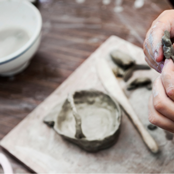 Photograph of hands shaping grey clay above a table where tools and clay are in place to make a ceramic bowl.